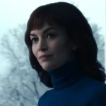 A scene from Severance, season 2, episode 9. Helly Eagan is shown in a bright setting, wearing a blue turtleneck sweater and displaying a contemplative expression. Behind her, a large window reveals a snowy landscape, contrasting with her calm yet meaningful gaze.