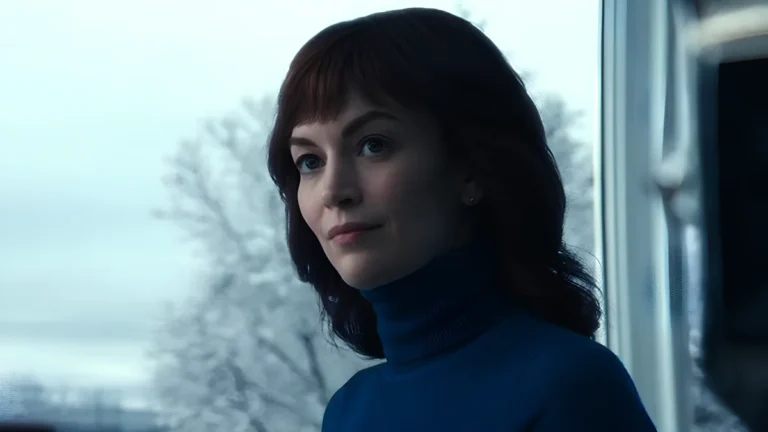 A scene from Severance, season 2, episode 9. Helly Eagan is shown in a bright setting, wearing a blue turtleneck sweater and displaying a contemplative expression. Behind her, a large window reveals a snowy landscape, contrasting with her calm yet meaningful gaze.