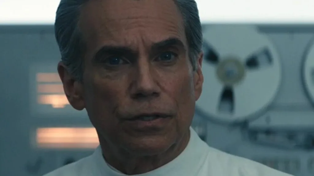 Dr. Mau, wearing a white coat, watches attentively with a rigid expression. In the background, recording tape reels spin in a machine.
