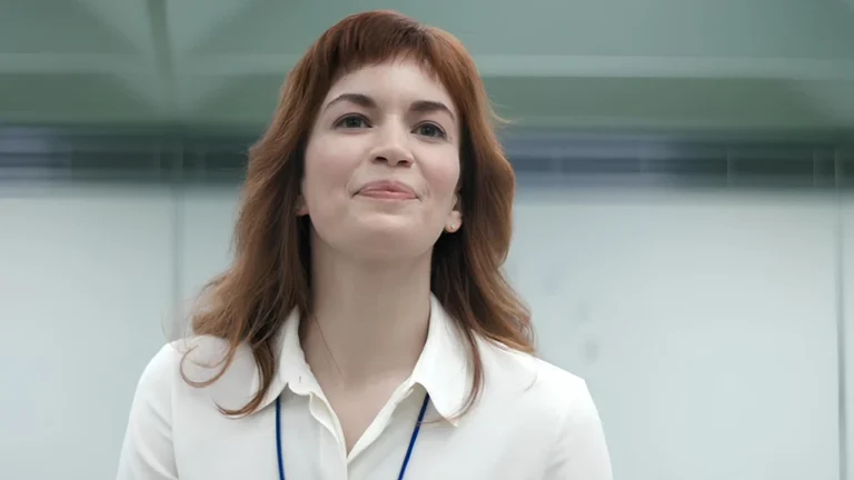 Helena Eagan in Severance gently smiling during her first day at Lumon's macrodata refinement department, wearing a white uniform and visible badge.