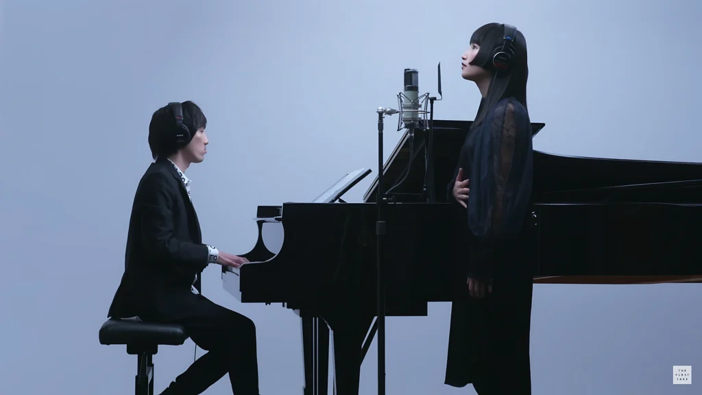 Hiroyuki Sawano tocando piano ao lado da cantora Eliana em um estúdio minimalista, durante apresentação para o canal The First Take.