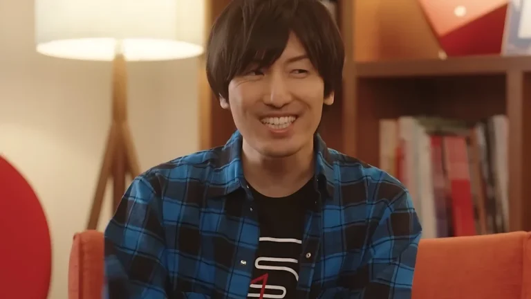 Hiroyuki Sawano smiling during a Netflix interview, wearing a blue plaid shirt in a relaxed setting with books in the background.