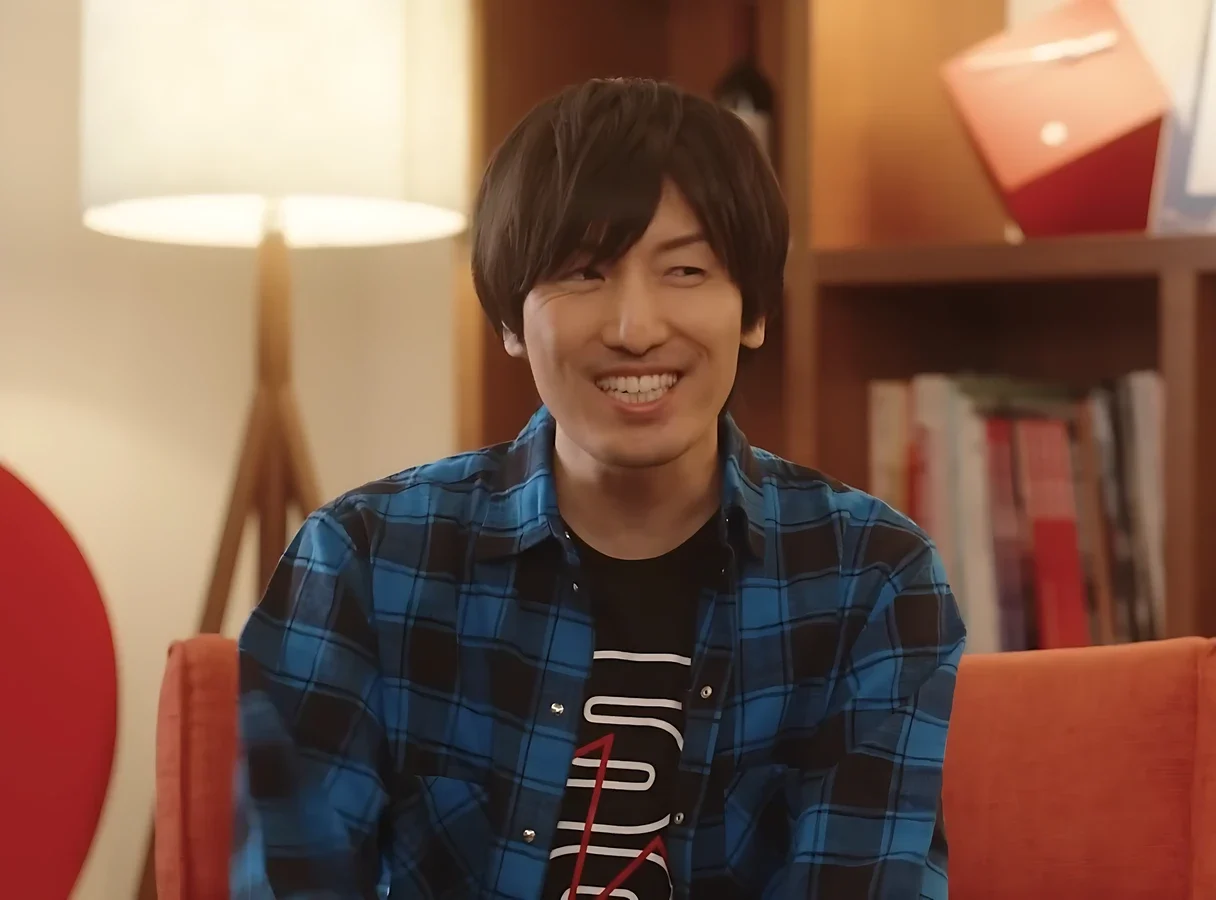Hiroyuki Sawano smiling during a Netflix interview, wearing a blue plaid shirt in a relaxed setting with books in the background.
