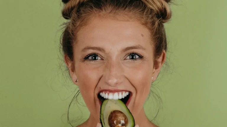 Woman with hair in buns holding and biting an avocado with a joyful expression, symbolizing healthy eating habits.
