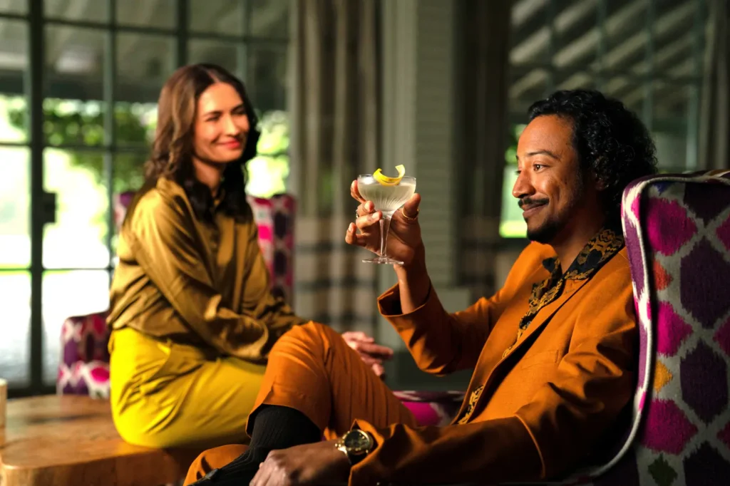 Koumba and Carol in Pluribus, seated in a sophisticated setting, representing the contrast between luxury, pleasure, and the moral collapse of the infected world.